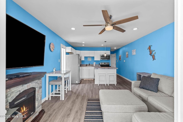 living area featuring a stone fireplace, light wood-style flooring, a ceiling fan, baseboards, and recessed lighting