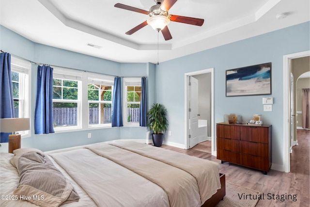 bedroom featuring a tray ceiling, wood finished floors, baseboards, ensuite bath, and a ceiling fan