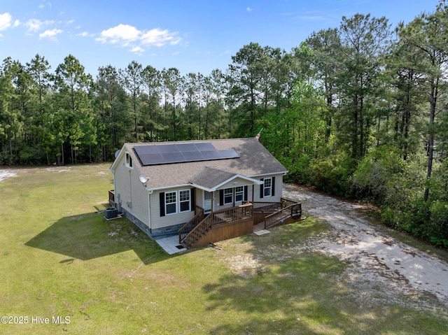 view of front facade featuring a front lawn, solar panels, a shingled roof, a deck, and a forest view