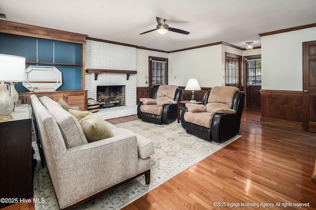 living area with wood finished floors, a wainscoted wall, ceiling fan, a brick fireplace, and crown molding
