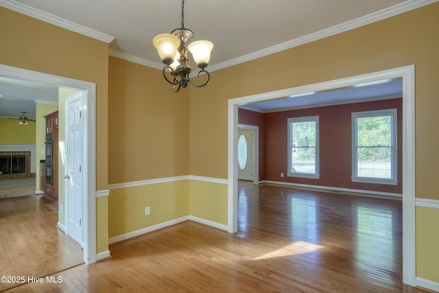 unfurnished room featuring crown molding, a brick fireplace, wood finished floors, baseboards, and ceiling fan with notable chandelier