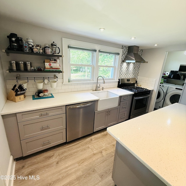 kitchen with stainless steel appliances, a sink, independent washer and dryer, wall chimney exhaust hood, and gray cabinetry