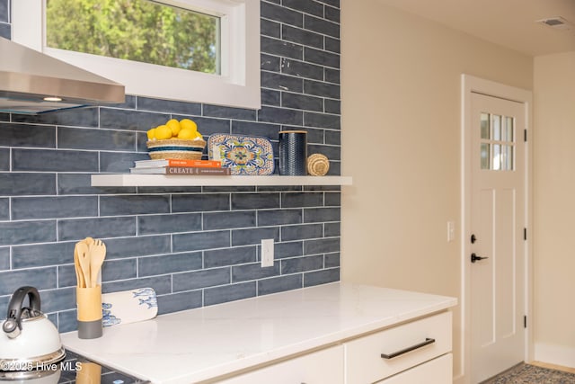 details featuring light stone counters, wall chimney range hood, white cabinets, backsplash, and visible vents