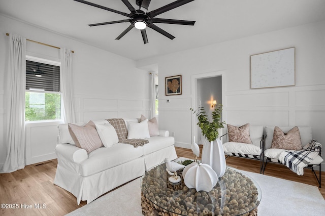living area featuring light wood-style floors, a decorative wall, ceiling fan, and a wainscoted wall