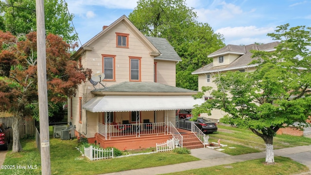 victorian-style house with a porch, fence, central AC unit, roof with shingles, and a front lawn