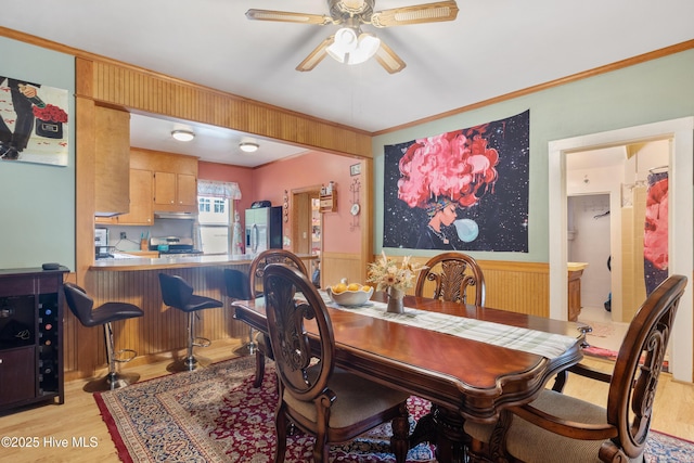 dining room featuring a wainscoted wall, a ceiling fan, ornamental molding, and light wood finished floors