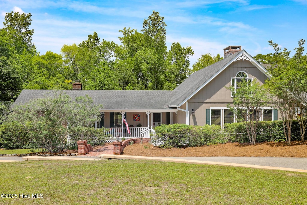 view of front of house featuring covered porch, a chimney, a shingled roof, and a front yard