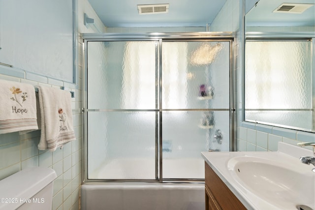 bathroom with visible vents, vanity, toilet, tile walls, and bath / shower combo with glass door