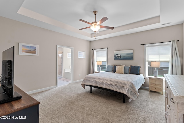 bedroom featuring a tray ceiling, baseboards, light carpet, ensuite bath, and ceiling fan