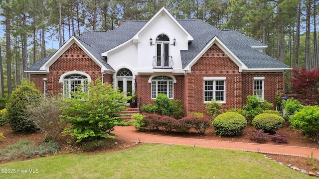 view of front facade with brick siding, roof with shingles, a balcony, and a front yard