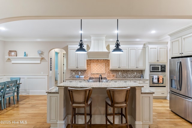 kitchen featuring appliances with stainless steel finishes, arched walkways, light wood-type flooring, a kitchen island with sink, and light stone counters
