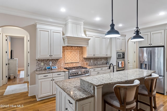kitchen featuring stainless steel appliances, arched walkways, premium range hood, an island with sink, and decorative backsplash