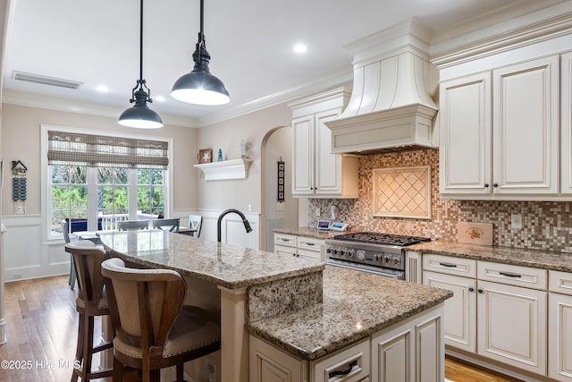 kitchen featuring a center island with sink, premium range hood, stainless steel range, light wood-style flooring, and light stone countertops