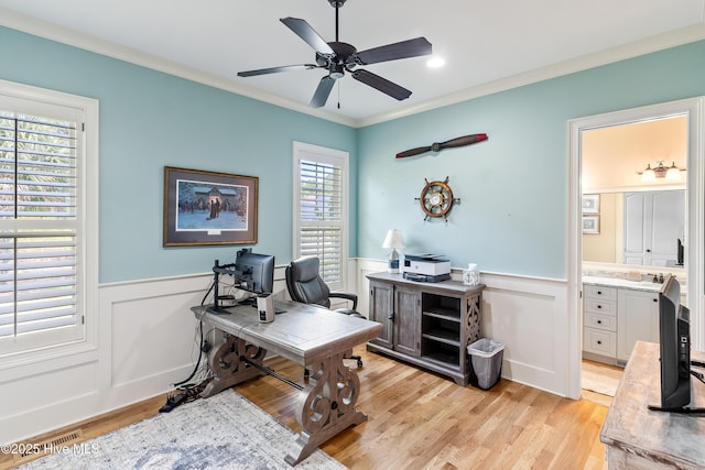 office area featuring wainscoting, crown molding, a ceiling fan, light wood-style flooring, and a decorative wall