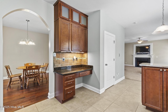 kitchen with a fireplace, glass insert cabinets, dark stone counters, tasteful backsplash, and ceiling fan with notable chandelier