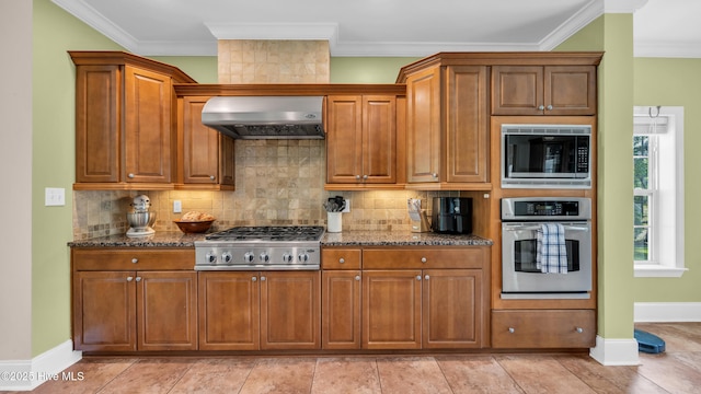 kitchen featuring stainless steel appliances, brown cabinetry, wall chimney exhaust hood, and dark stone countertops