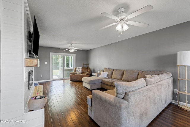 living room featuring baseboards, dark wood-style floors, a textured ceiling, a large fireplace, and a ceiling fan