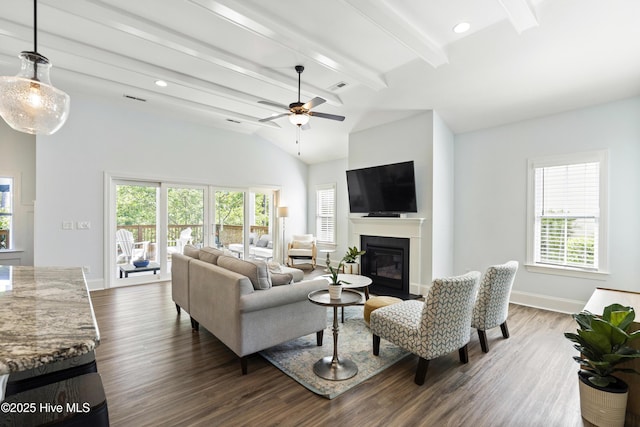 living area with ceiling fan, baseboards, dark wood-style floors, a glass covered fireplace, and vaulted ceiling with beams