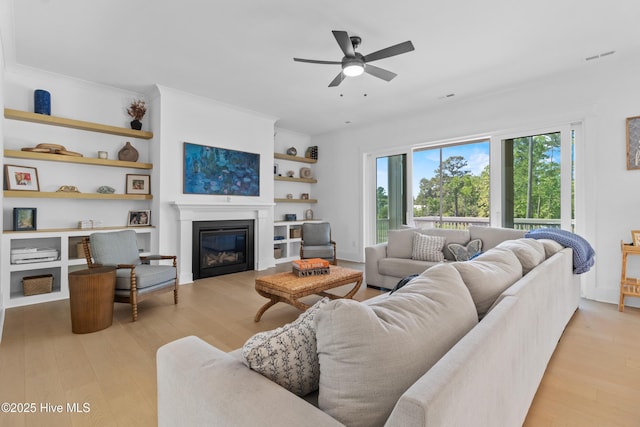 living area featuring ceiling fan, visible vents, light wood-type flooring, built in shelves, and a glass covered fireplace