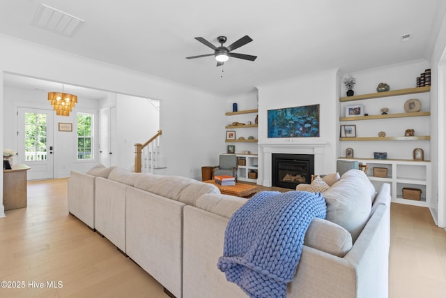 living area with a glass covered fireplace, built in features, ceiling fan with notable chandelier, light wood-type flooring, and visible vents