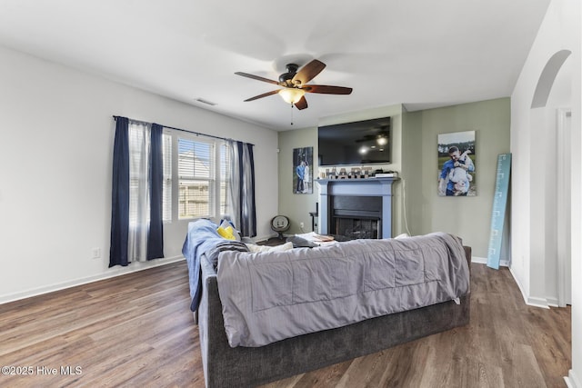 bedroom with a ceiling fan, visible vents, a fireplace, wood finished floors, and baseboards