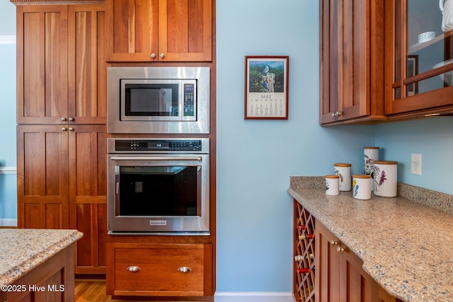 kitchen featuring light stone countertops, brown cabinetry, glass insert cabinets, and appliances with stainless steel finishes