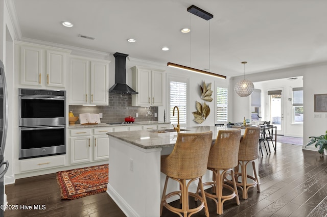 kitchen with a sink, double oven, dark wood finished floors, wall chimney exhaust hood, and white cabinetry