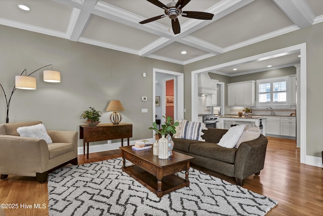 living room with a ceiling fan, coffered ceiling, light wood finished floors, beamed ceiling, and ornamental molding