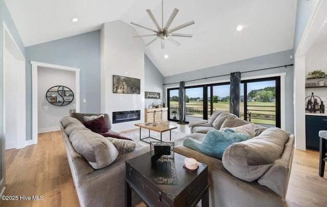 living room featuring a ceiling fan, light wood-type flooring, high vaulted ceiling, baseboards, and a glass covered fireplace