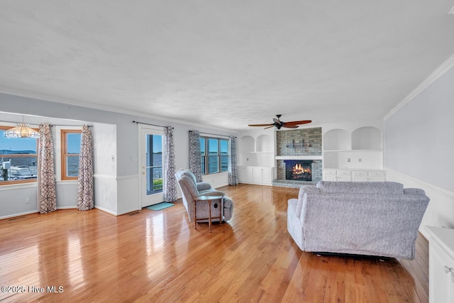 living area with a ceiling fan, crown molding, a fireplace, built in shelves, and light wood-style flooring