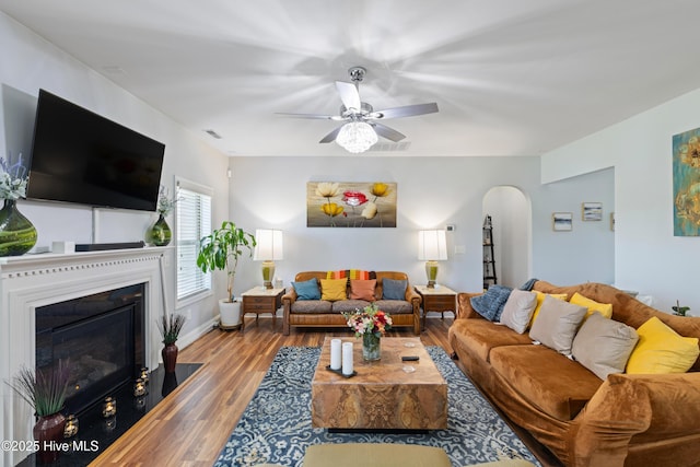 living room with wood finished floors, arched walkways, a fireplace with flush hearth, and a ceiling fan
