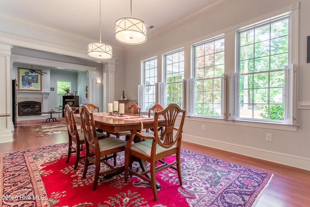 dining space featuring a fireplace with raised hearth, baseboards, wood finished floors, ornate columns, and ornamental molding