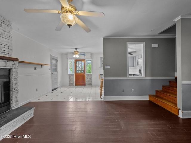 unfurnished living room featuring crown molding, ceiling fan, a stone fireplace, stairs, and baseboards
