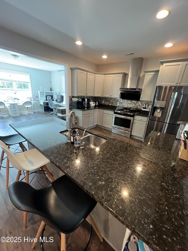 kitchen featuring dark stone counters, wall chimney exhaust hood, a sink, backsplash, and stainless steel appliances
