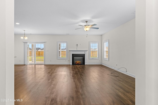 unfurnished living room featuring baseboards, a warm lit fireplace, ceiling fan with notable chandelier, and dark wood-style floors
