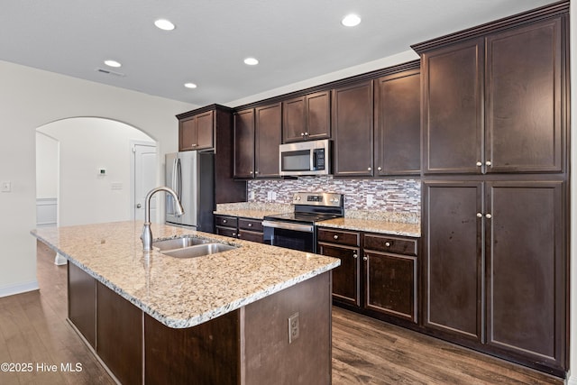 kitchen featuring arched walkways, dark wood-style flooring, an island with sink, and stainless steel appliances