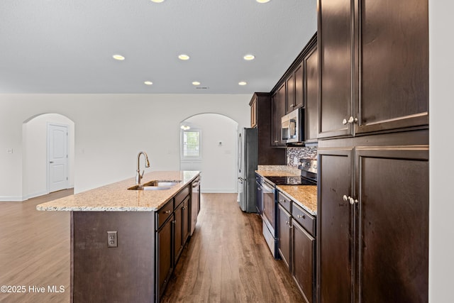 kitchen featuring a kitchen island with sink, dark brown cabinetry, arched walkways, appliances with stainless steel finishes, and a sink