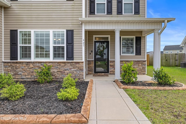 doorway to property with fence, stone siding, cooling unit, a lawn, and covered porch