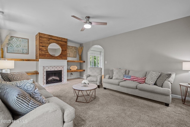 carpeted living area featuring ceiling fan, arched walkways, baseboards, a tile fireplace, and visible vents