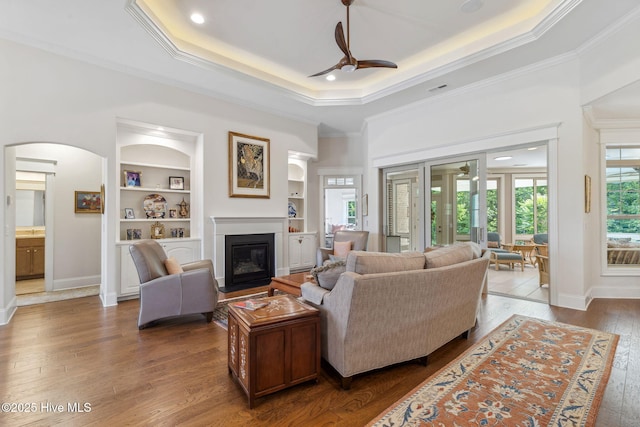 living room with a ceiling fan, hardwood / wood-style floors, arched walkways, a glass covered fireplace, and built in shelves