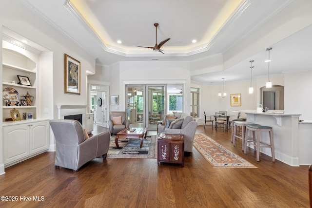 living area with a fireplace, a raised ceiling, built in shelves, ornamental molding, and dark wood-style floors