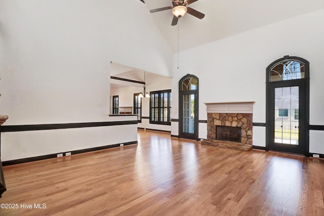 unfurnished living room with a towering ceiling, wood finished floors, a stone fireplace, baseboards, and ceiling fan