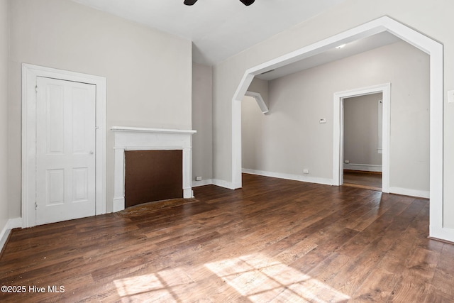 unfurnished living room featuring ceiling fan, arched walkways, baseboards, a fireplace with flush hearth, and wood finished floors