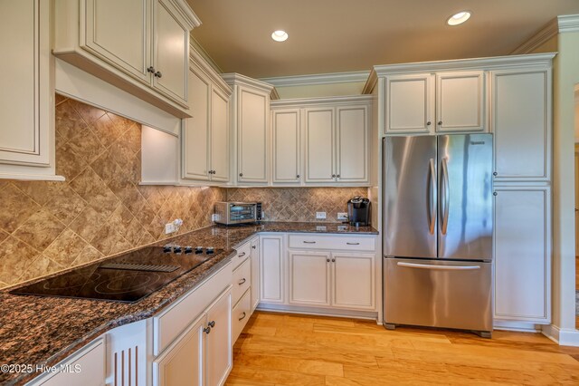 kitchen featuring light wood-style floors, tasteful backsplash, black electric stovetop, stainless steel fridge, and dark stone counters