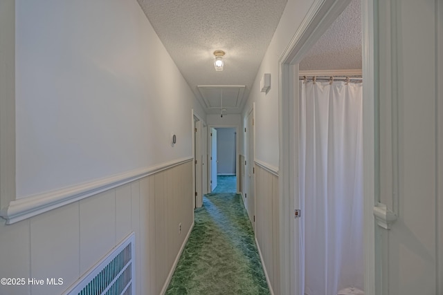 corridor featuring attic access, a textured ceiling, visible vents, carpet floors, and a wainscoted wall