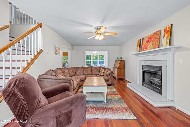 living area with a ceiling fan, wood finished floors, a fireplace with raised hearth, and stairs