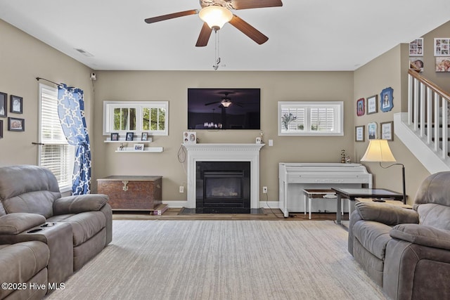 living area featuring a glass covered fireplace, a ceiling fan, wood finished floors, visible vents, and stairway