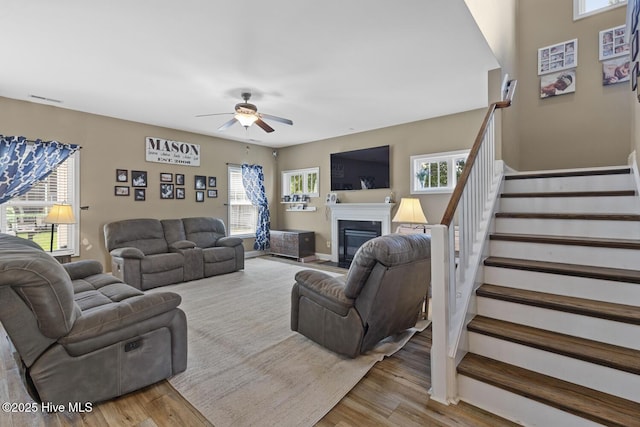 living area featuring a ceiling fan, visible vents, a fireplace with flush hearth, wood finished floors, and stairs