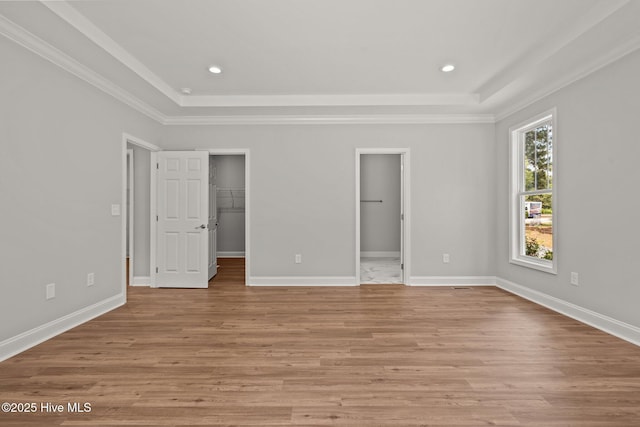 unfurnished bedroom featuring a raised ceiling, a spacious closet, light wood finished floors, crown molding, and baseboards