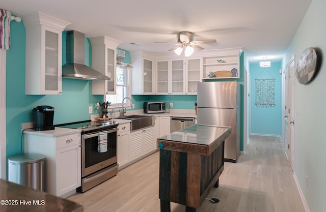 kitchen featuring appliances with stainless steel finishes, wall chimney range hood, a sink, white cabinetry, and a ceiling fan